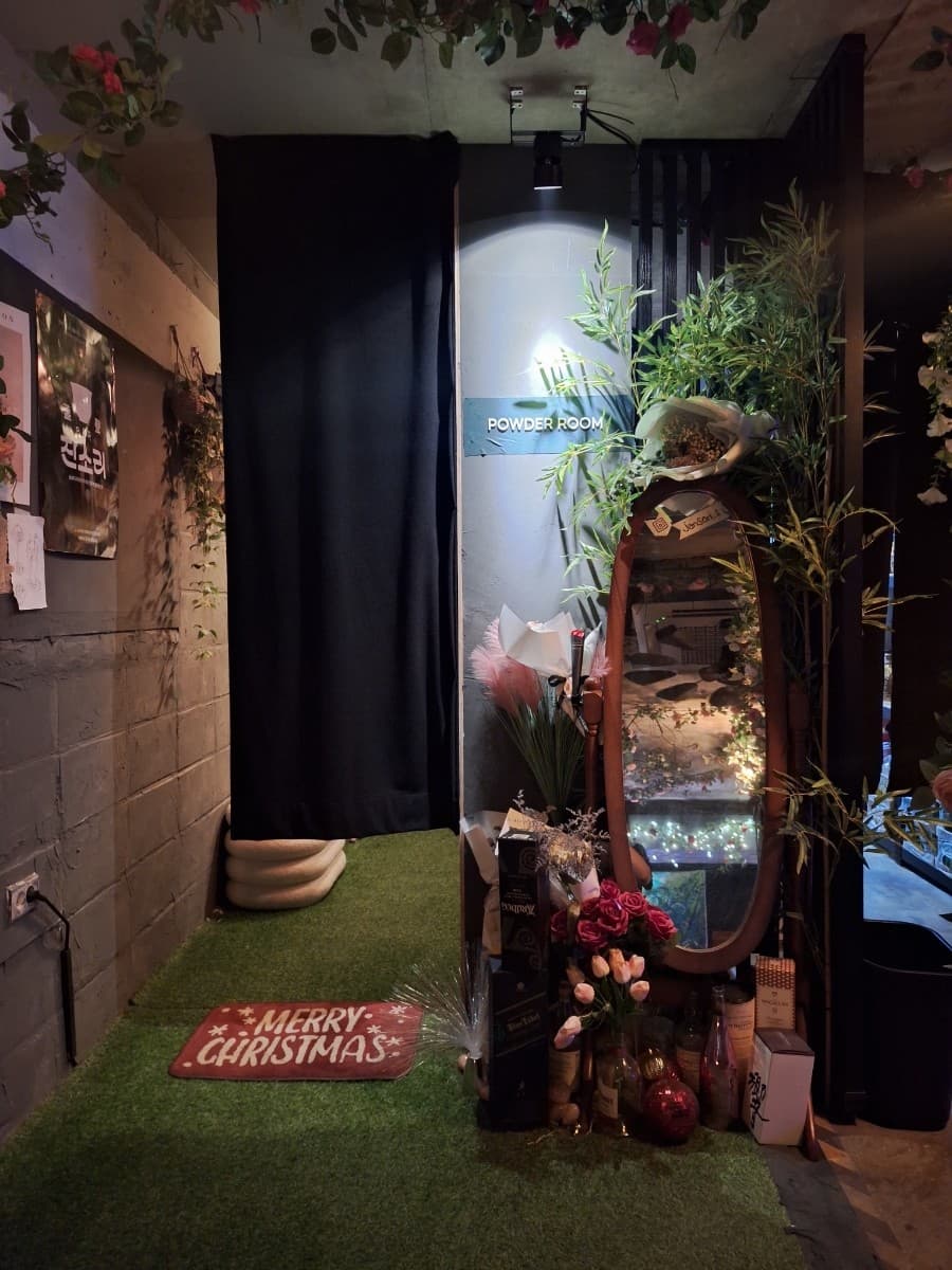 대전광역시 유성구 혼술바잔소리 봉명점 대전 바(Bar)의 핀 조명이 비추는 전신 거울과 꽃 장식. 인조 잔디와 위스키 병으로 꾸며진 파우더룸이 1인 혼술객을 위한 동선에 맞춰 배치됨.
