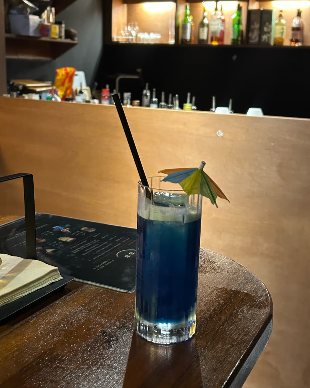 대전광역시 중구 혼술바히히 대전 바(Bar)의 나무 다찌석 위 파란색 칵테일과 종이 우산 장식. 1인 혼술을 위한 좌석에서 보이는 배경의 술병 선반과 노란 조명.