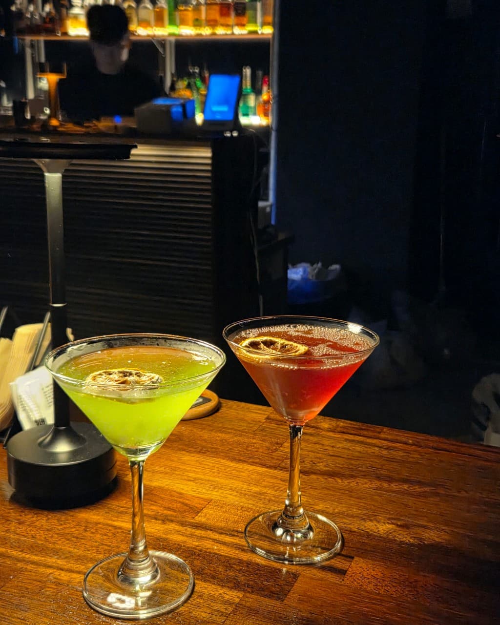 대전광역시 중구 노웨이브 대전혼술바 대흥점 바(Bar) 나무 다찌석 위 조명을 받는 연두색과 붉은색 칵테일. 말린 레몬 칩이 띄워진 잔과 어두운 실내 배경의 1인 카운터석에서 즐기는 혼술 상황.
