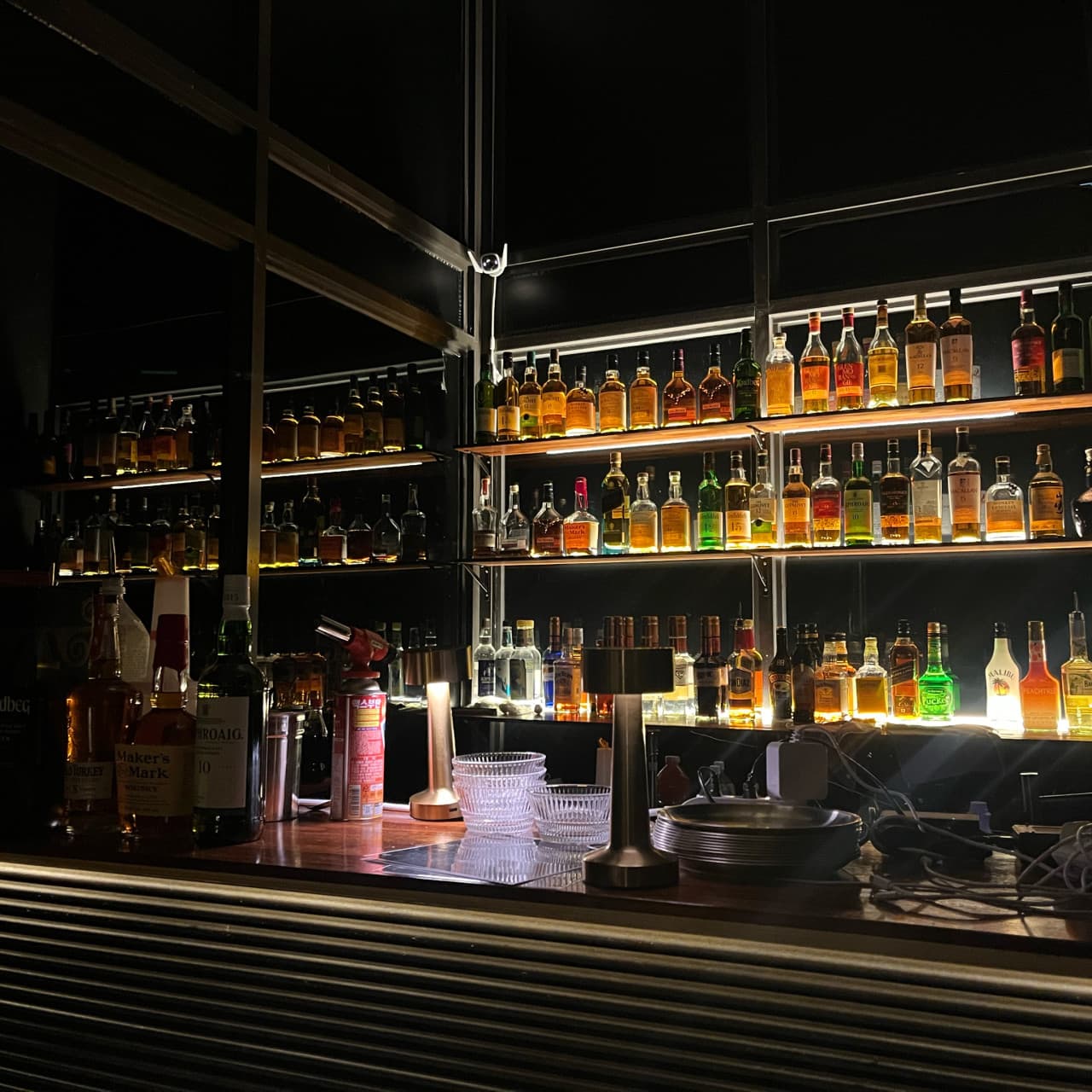 대전광역시 중구 노웨이브 대전혼술바 대흥점 바(Bar)의 조명 비치는 위스키 진열장과 나무 다찌석. 1인 혼술을 위해 마련된 카운터 테이블 위 유리잔과 바 도구.