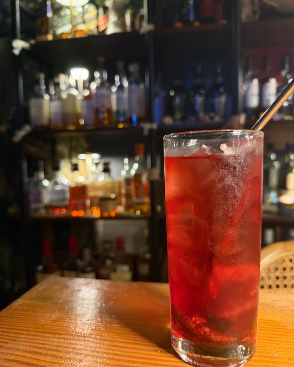 대전광역시 서구 혼술바프시케 대전 바(Bar) 다찌석에 놓인 얼음 가득한 붉은 칵테일. 나무 테이블 위 금속 빨대와 조명 받은 술병 진열장 배경의 1인 혼술 상황.