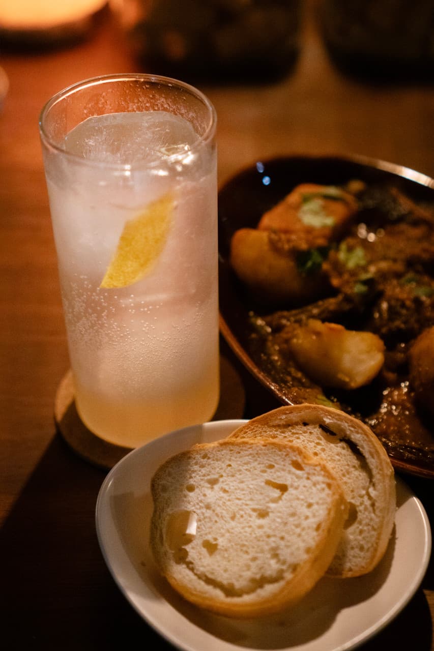 대전광역시 서구 탱이 대전 바(Bar) 다찌석 혼술. 탄산 기포가 맺힌 레몬 하이볼과 바게트, 진한 소스의 고기 요리. 노란 조명 아래 차려진 1인 상차림.