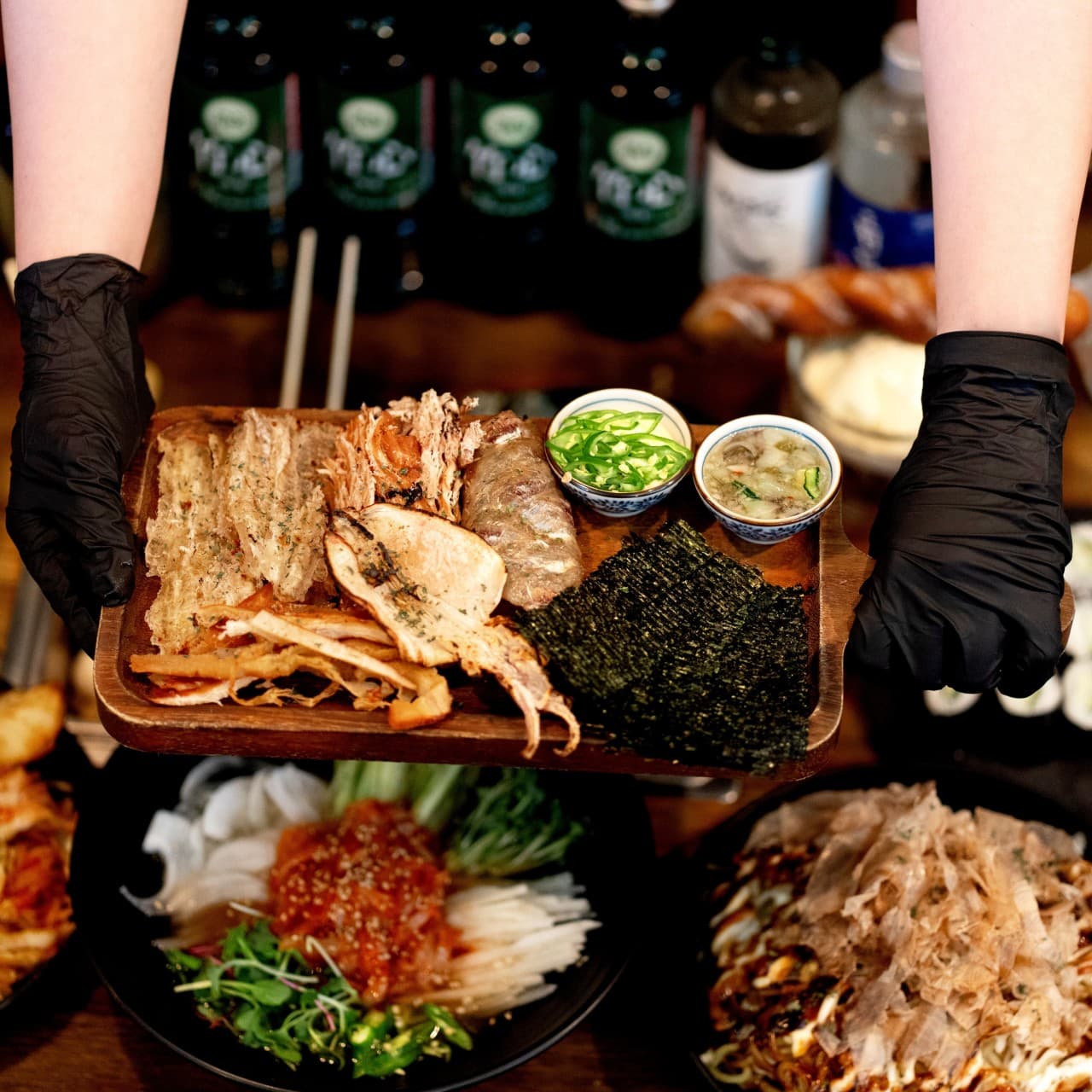 대전광역시 서구 요리주점 철길부산집 가수원점 대전 다찌석 1인 혼술. 검은 장갑을 낀 손이 건어물, 김, 소스가 담긴 나무 트레이를 서빙하며 조명 아래 안주의 질감을 보여줌.