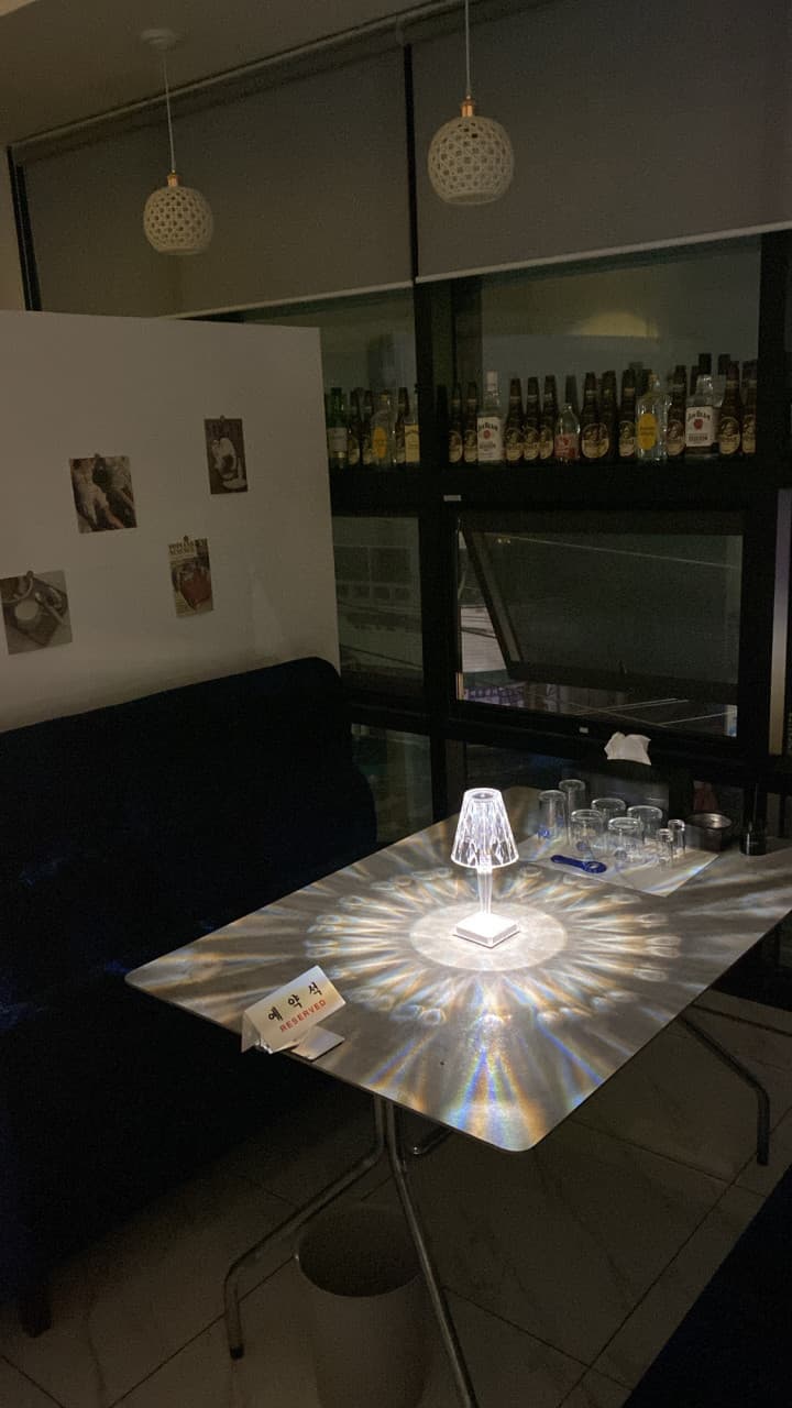 경상북도 칠곡군 여우bar 경북 바(Bar). 기하학적 빛 패턴의 테이블 조명이 비추는 1인 소파 좌석과 예약석 팻말. 창가에 나열된 술병들과 어두운 실내에서 즐기는 혼술 상황.