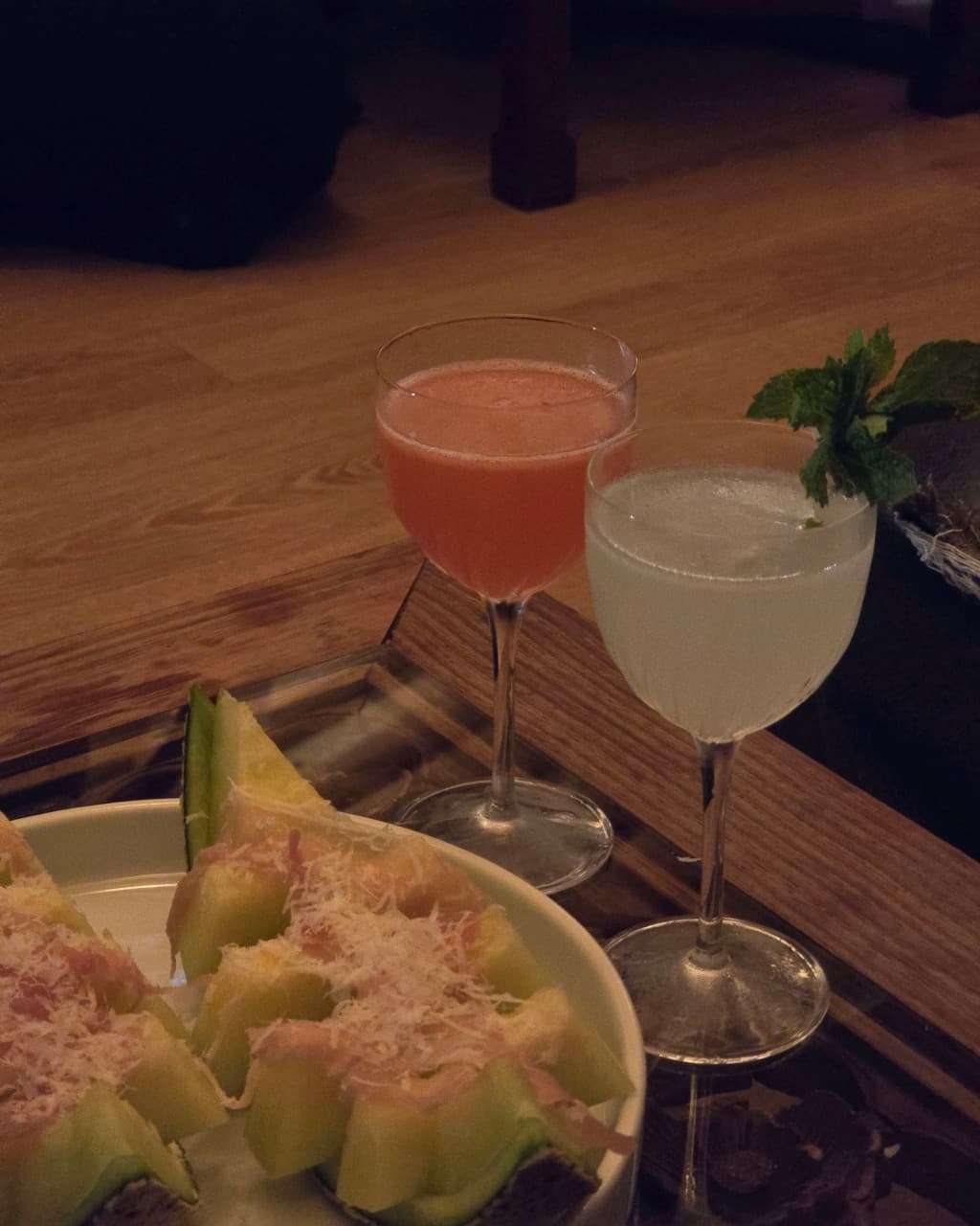 경상북도 경주시 바분 월정교점 경북 바(Bar) 혼술. 낮은 조명 아래 나무 테이블 위 치즈 가루를 뿌린 멜론 하몽 안주와 두 잔의 칵테일이 놓인 다찌석.