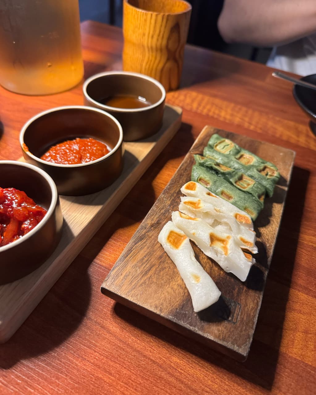 경기도 수원시 매산약수터 수원역본점 경기 요리주점 - 주황빛 조명 아래 나무 도마 위 노릇한 가래떡과 소스 3종이 차려진 1인 혼술 테이블