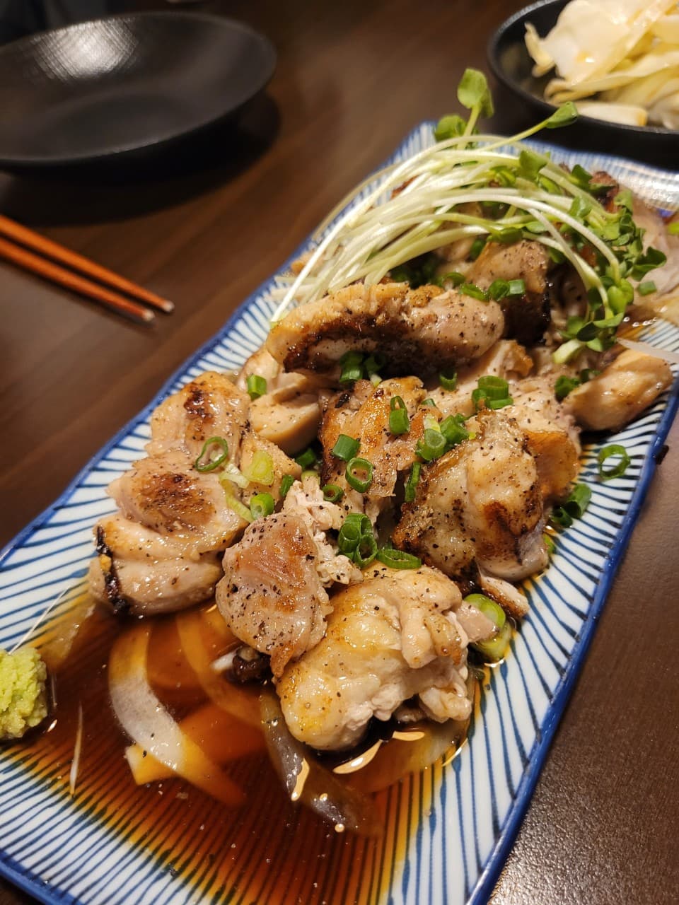 경기도 부천시 이자카야 철판오사카야 부천점 경기 다찌석 조명 아래 차려진 1인 혼술 안주. 소스가 배어든 닭구이와 무순, 와사비가 담긴 접시의 질감을 묘사함.