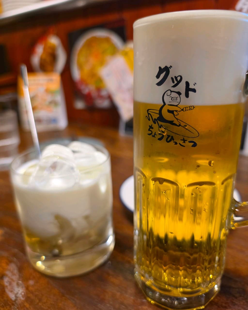 부산광역시 수영구 초힛사츠 부산 이자카야 나무 다찌석. 캐릭터 잔의 생맥주 거품과 얼음 담긴 술이 놓인 혼술 상황. 조명이 비치는 목재 테이블 위 주류의 질감.
