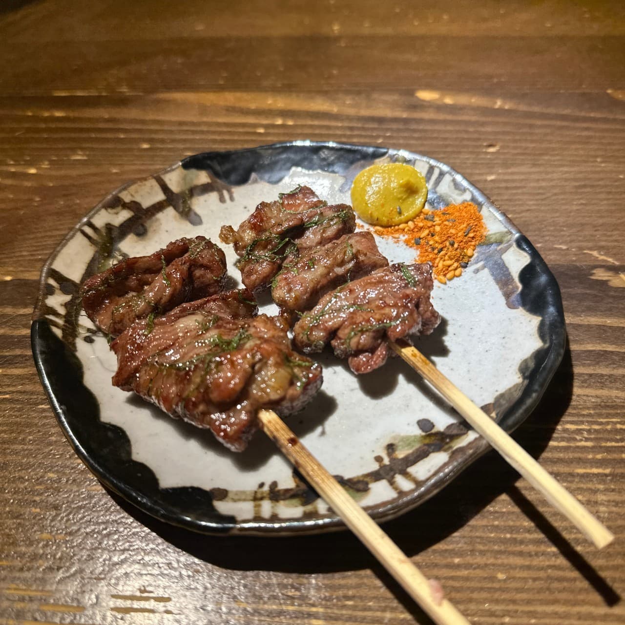 부산광역시 수영구 이자카야 초힛사츠 부산 나무 다찌석에서 혼술과 곁들이는 윤기 흐르는 꼬치구이. 어두운 배경 속 조명이 비추는 접시 위 겨자와 시치미 가루.