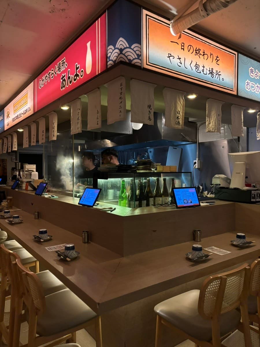 부산광역시 부산진구 앙요 전포본점 부산 요리주점. 나무 다찌석과 1인 식기, 태블릿이 구비된 혼술 공간. 상단 조명 아래 주방의 조리 현장과 진열된 주류가 보이는 구성.