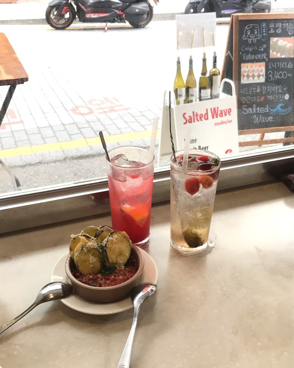 부산광역시 부산진구 솔티드웨이브 전포 부산 와인바 창가 다찌석 1인 테이블. 자연광 아래 치즈 가루를 뿌린 가지 요리와 과일이 담긴 와인 두 잔이 놓인 혼술 상황.