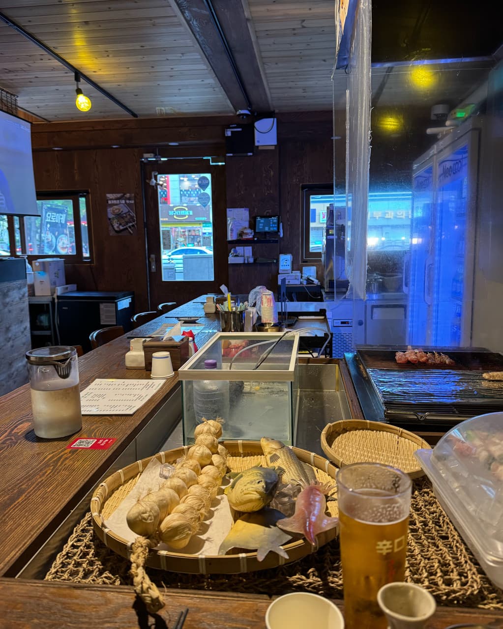 대구광역시 중구 요로이케 교동점 이자카야 - 노란 조명 아래 나무 다찌석과 생맥주, 꼬치구이 화로가 배치된 1인 혼술 전용 공간