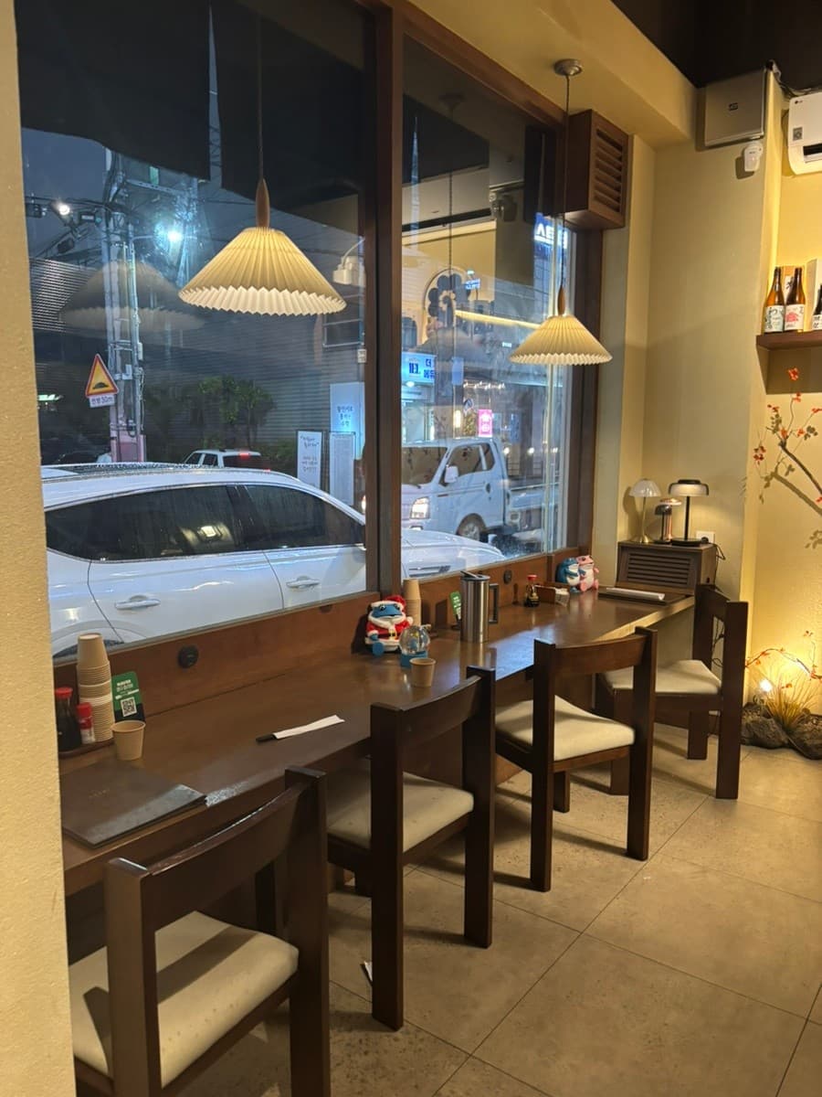 대구광역시 수성구 이자카야 와코안 수성범어점 대구의 주름 갓 조명 아래 창가 다찌석 1인 좌석. 나무 테이블 위 메뉴판과 컵이 놓여 있고 유리창 밖으로 밤 거리가 보이는 혼술 식당.