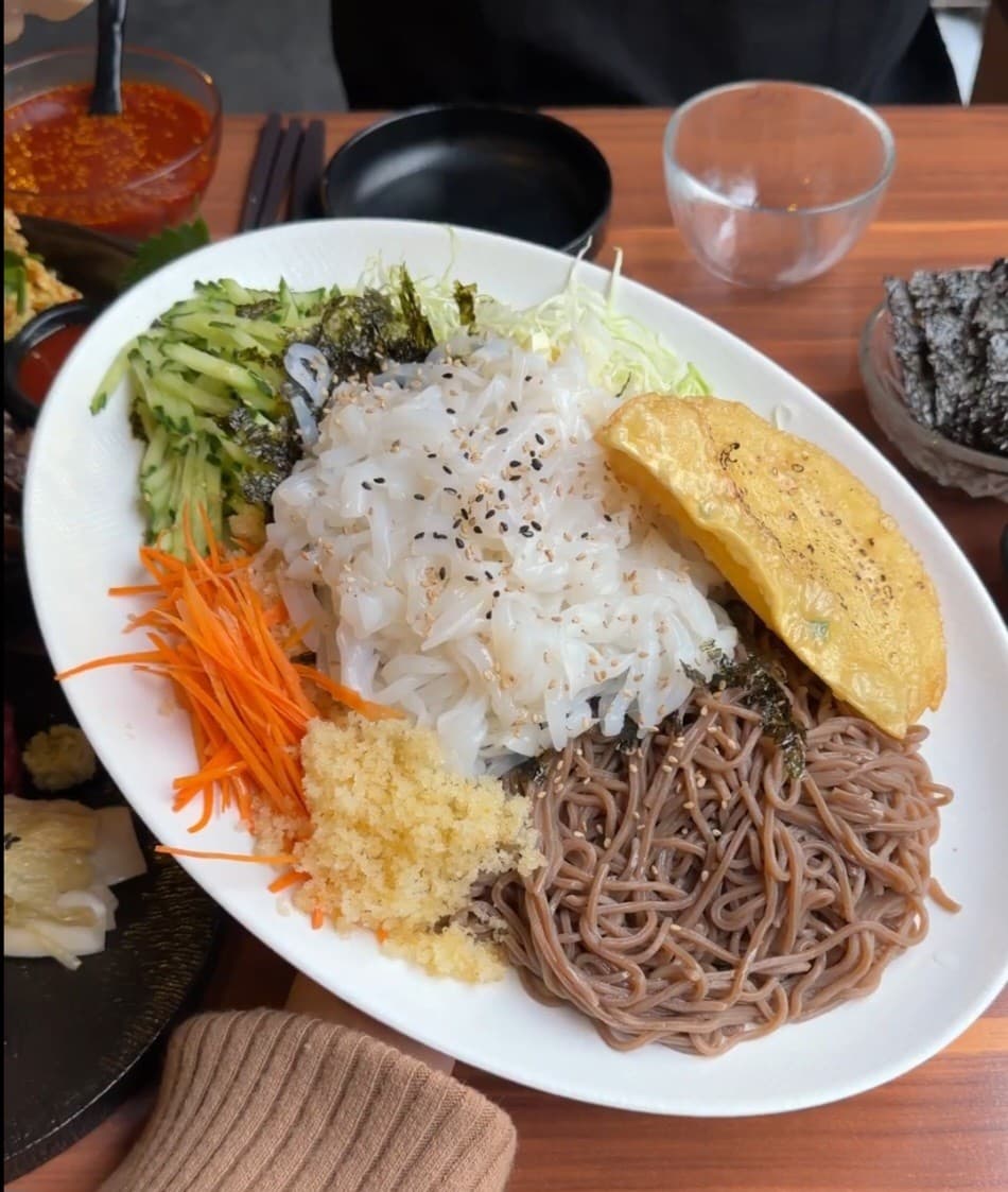부산광역시 동래구 월하주방 부산 요리주점. 나무 테이블 위 1인 다찌석에 차려진 오징어회와 메밀면, 채소 고명. 조명 아래서 혼자 식사와 술을 즐기는 상황.