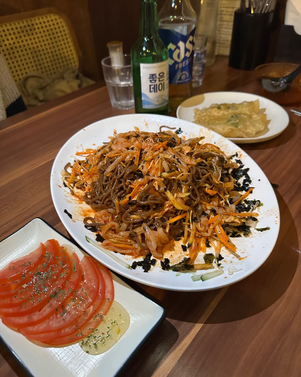 부산광역시 동래구 요리주점 월하주방 부산의 나무 테이블 위 빨간 양념의 비빔면과 토마토 슬라이스. 조명 아래 소주와 맥주가 차려진 1인 혼술 상황.