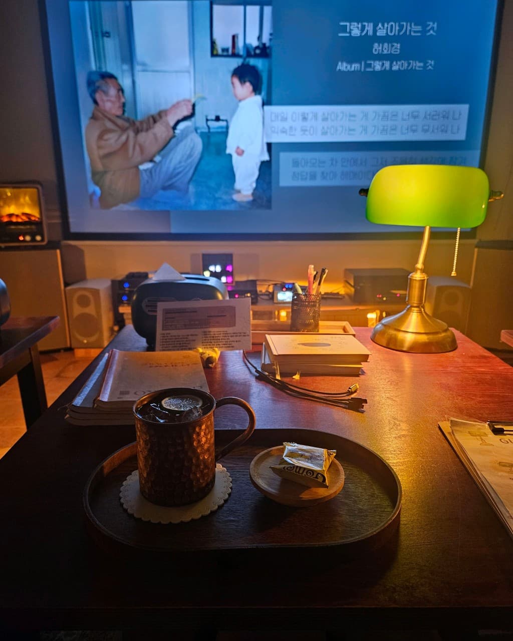 부산광역시 수영구 광안리골방 부산 바(Bar). 초록색 스탠드 조명이 켜진 1인석 테이블 위 구리잔 음료와 간식, 정면 스크린에 띄워진 음악 가사 영상을 응시하는 혼술 현장.