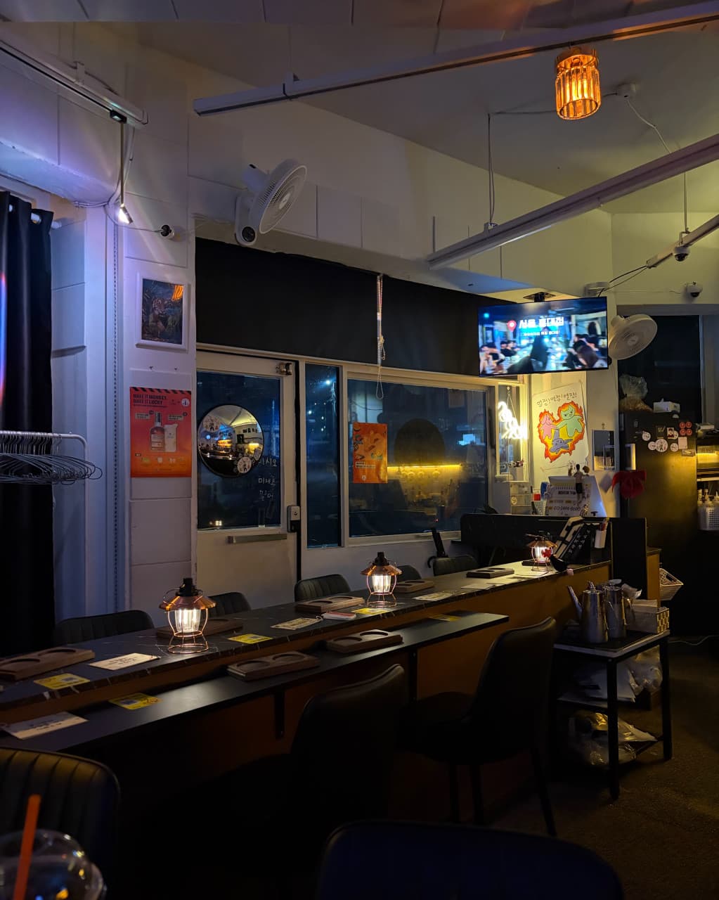 부산광역시 수영구 자유의지 혼술바 부산광안리점 바(Bar) 다찌석 위 램프 조명과 1인 좌석들. 벽면 포스터와 TV 화면이 배치된 낮은 조도의 공간에서 혼술을 즐기는 상황.