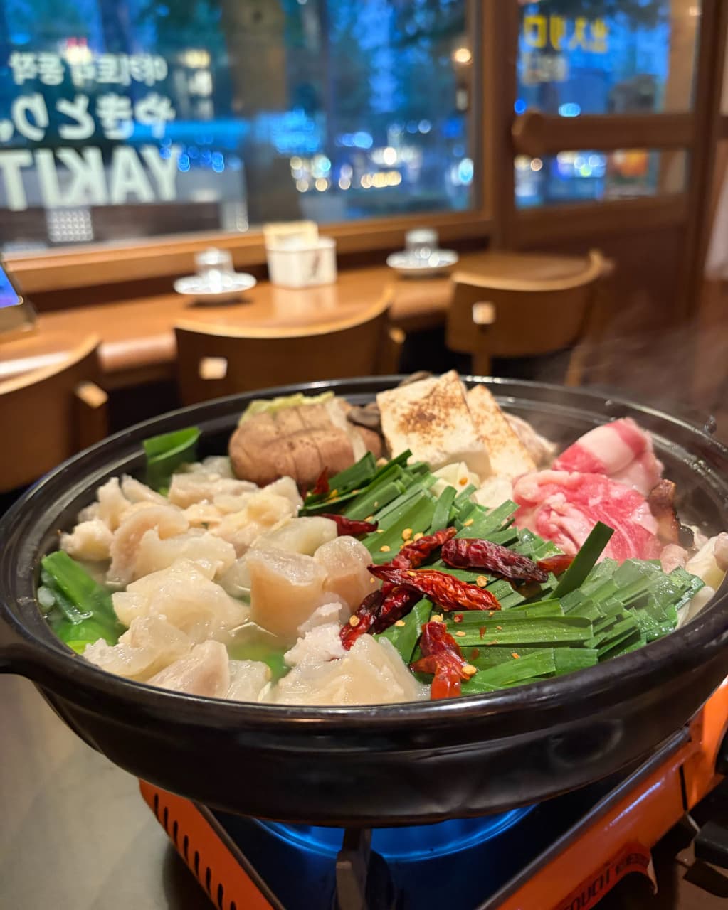 인천광역시 남동구 야키토리동작 인천시청점 이자카야의 다찌석. 1인 버너 위 대창과 부추가 담긴 전골의 질감과 김이 올라오는 상황. 창밖 조명 아래서 즐기는 1인 혼술 자리.