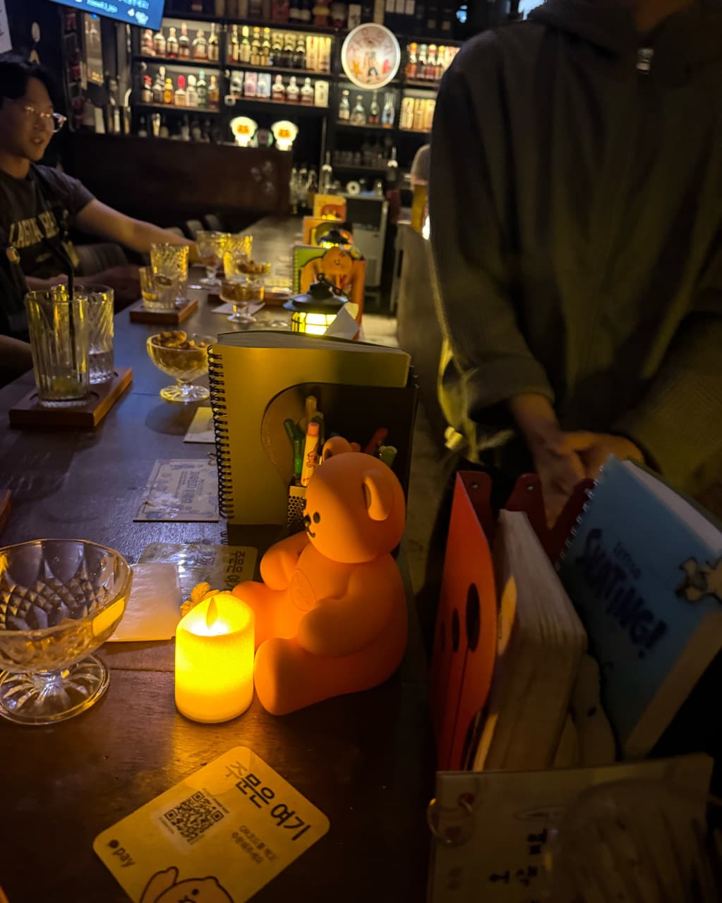 제주특별자치도 제주시 자유의지 혼술바 제주본점 바(Bar)의 다찌석. 노란 촛불 조명과 곰돌이 소품, 메뉴판이 놓인 1인 좌석 뒤로 술병이 진열된 혼술 현장.