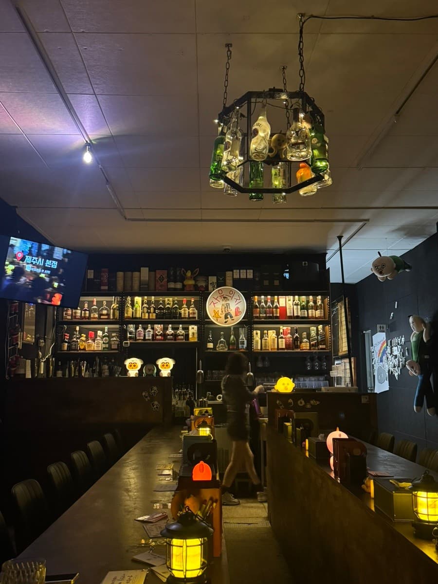 제주특별자치도 제주시 자유의지 혼술바 제주본점 바(Bar). 술병 샹들리에와 노란 랜턴 조명이 비치는 1인 다찌석에서 혼술을 즐기는 상황.