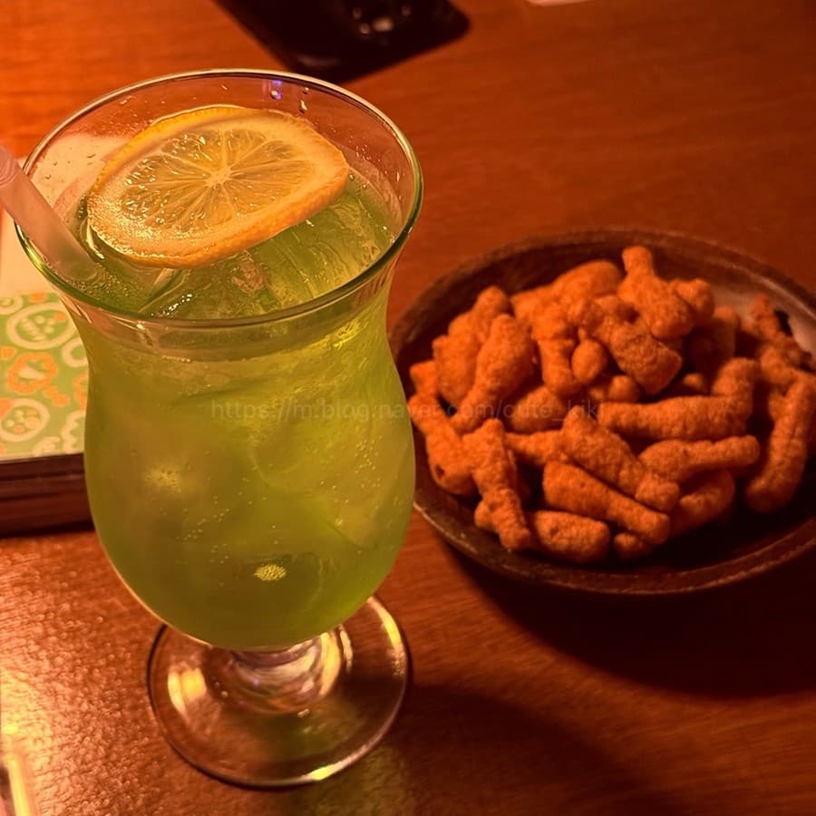 제주특별자치도 제주시 단지 제주혼술바 바(Bar)의 주황색 조명 아래 레몬을 띄운 초록색 하이볼과 닭다리 과자. 나무 테이블 1인 다찌석에서 혼술을 즐기는 상황.