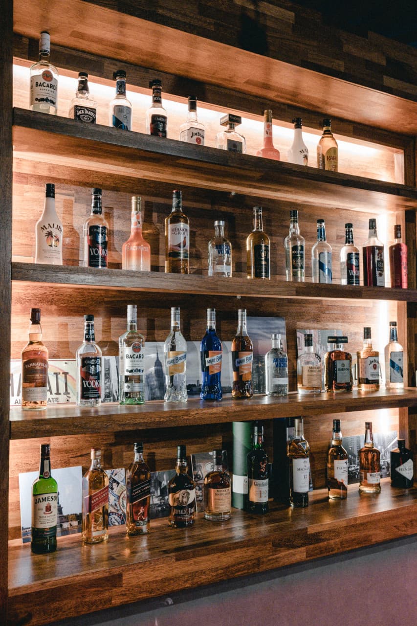 제주특별자치도 서귀포시 제주혼술바 언스테이블 서귀포점 바(Bar) - 원목 진열장에 정렬된 위스키와 보드카 병들 위로 비치는 노란 간접 조명과 1인 혼술 공간