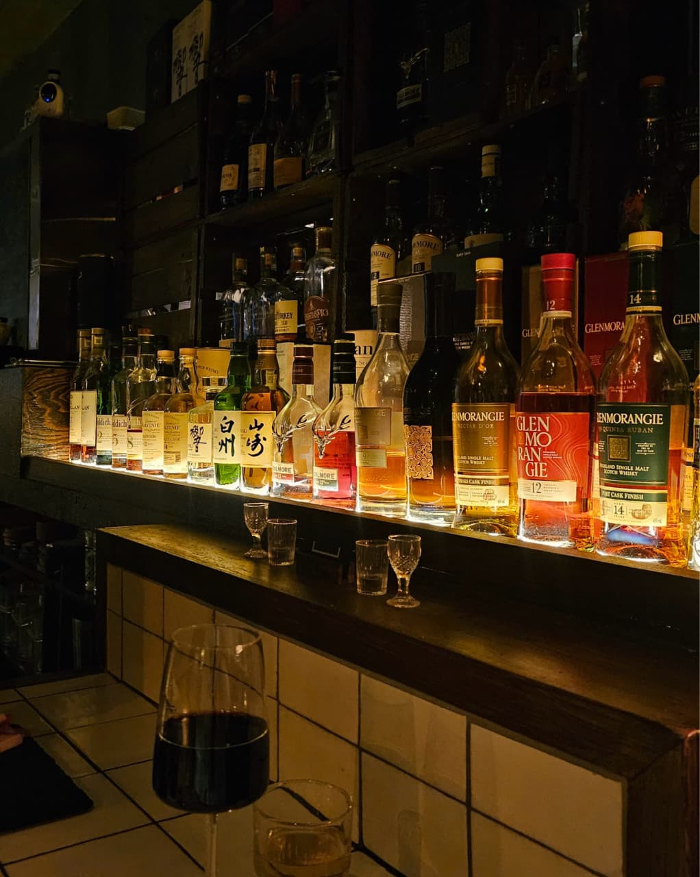 제주특별자치도 서귀포시 명단 BBQ BAR 제주 1인 다찌석. 조명 아래 위스키와 기타 주류 병이 진열된 타일 테이블 위 레드 와인이 놓인 혼술 상황.