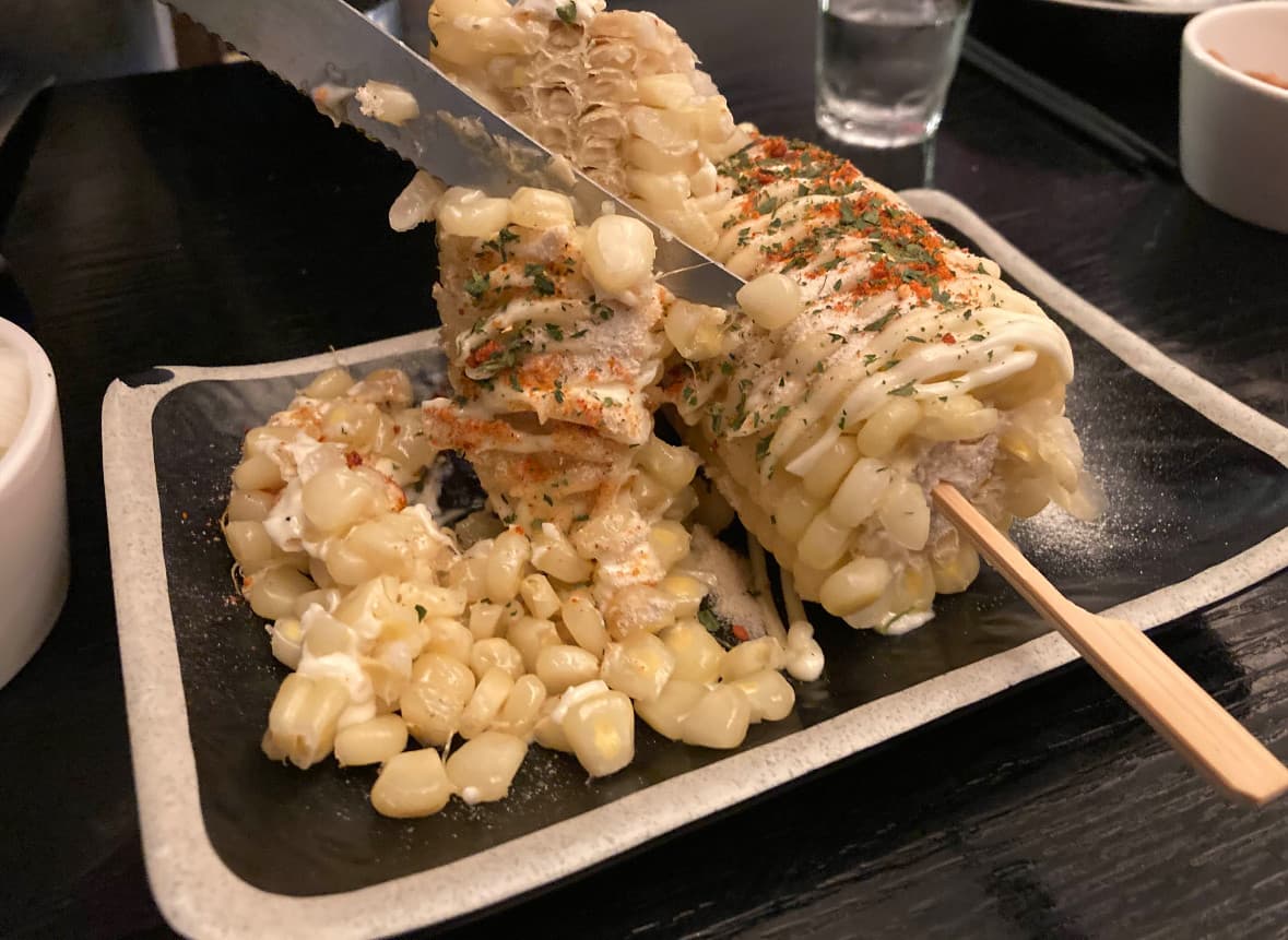 대전광역시 유성구 혼술 이자카야 꼬치는밤 대전. 어두운 테이블 위 다찌석에서 나이프로 마약옥수수 알갱이를 발라내는 상황과 시즈닝이 뿌려진 옥수수 질감.