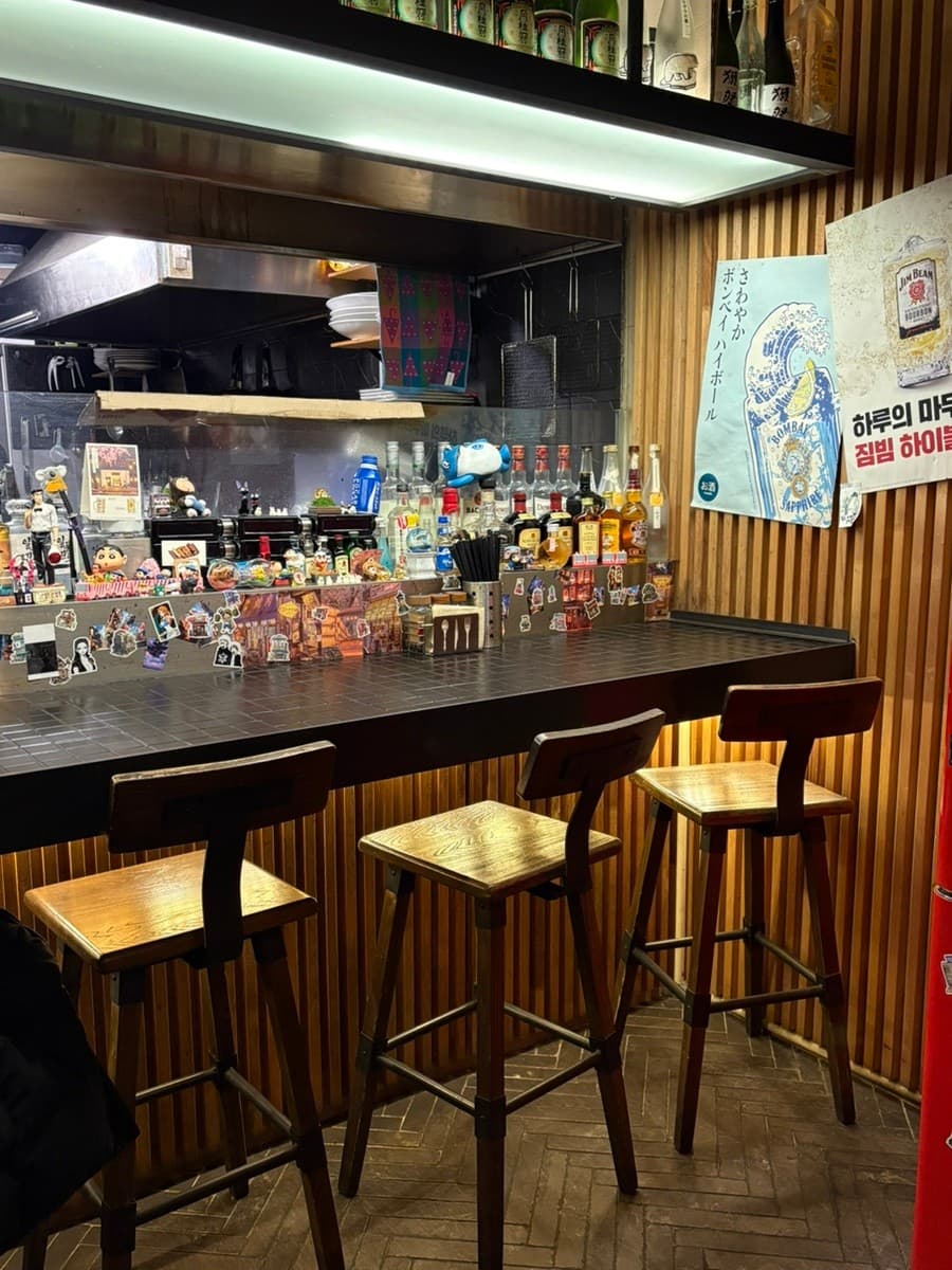 대전광역시 유성구 이자카야 꼬치는밤 대전의 1인 다찌석. 하이볼 포스터와 술병이 진열된 나무 벽면 아래 조명이 비치는 카운터 좌석이 배치된 혼술 공간.