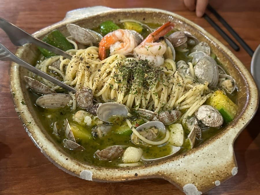 경기도 안산시 요리주점 반월가 경기. 나무 테이블 위 바지락과 새우를 곁들인 파스타가 놓인 1인 혼술 상황. 따뜻한 조명 아래 해산물과 면발의 질감이 강조됨.
