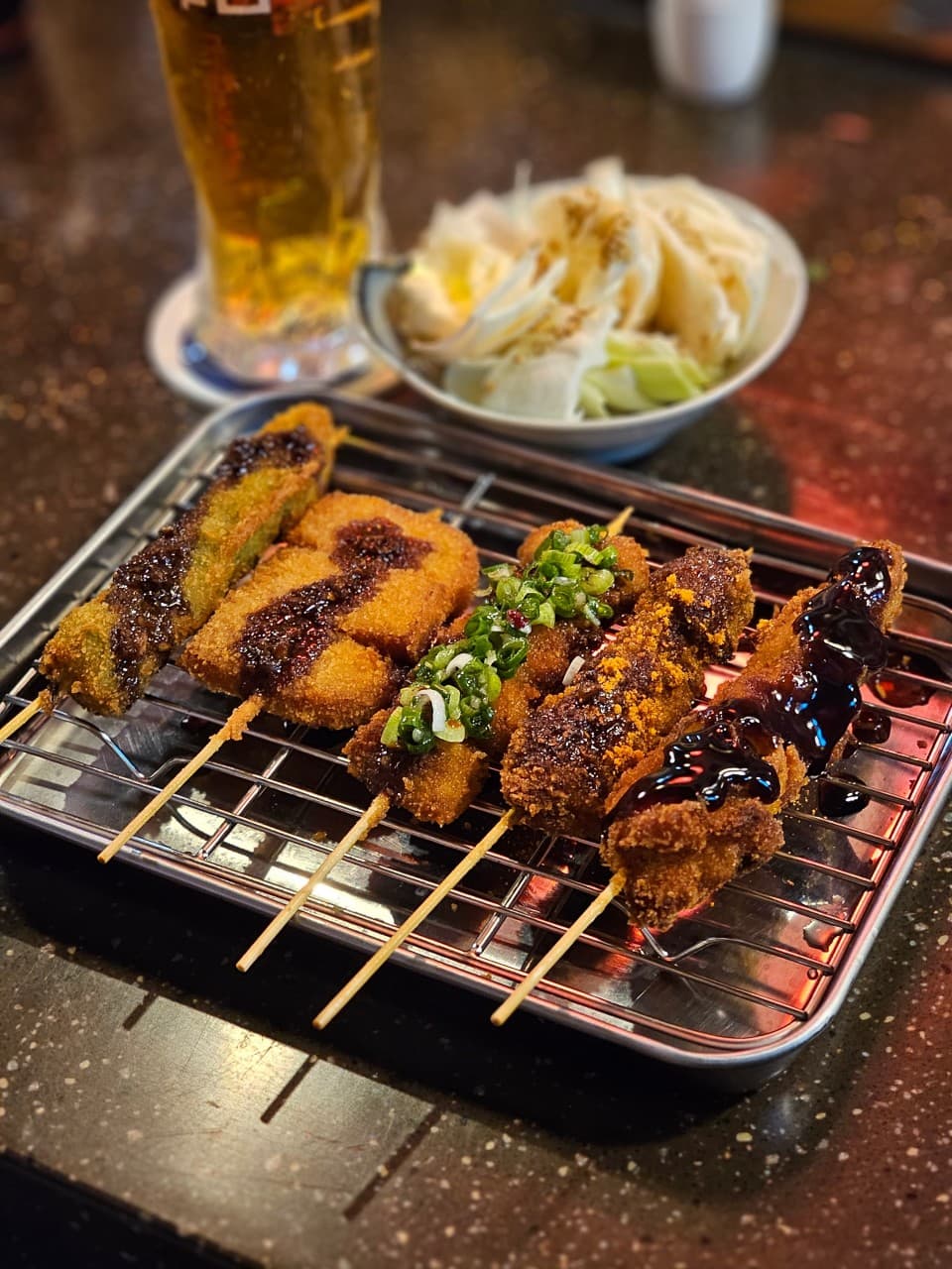 경기도 수원시 나츠비 율전동본점 경기 이자카야 다찌석 혼술. 소스를 얹은 바삭한 질감의 쿠시카츠 꼬치들과 양배추, 맥주가 어두운 조명 아래 놓인 상황.