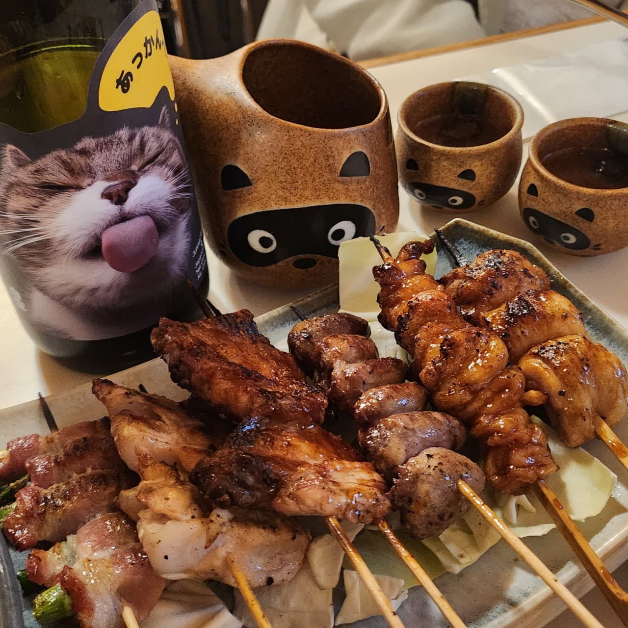 경기도 안양시 요리주점 마시타 경기 다찌석 1인 혼술. 양념이 발린 닭꼬치 구이와 고양이 라벨 술병, 너구리 모양 토기 술잔이 조명 아래 놓인 식탁.