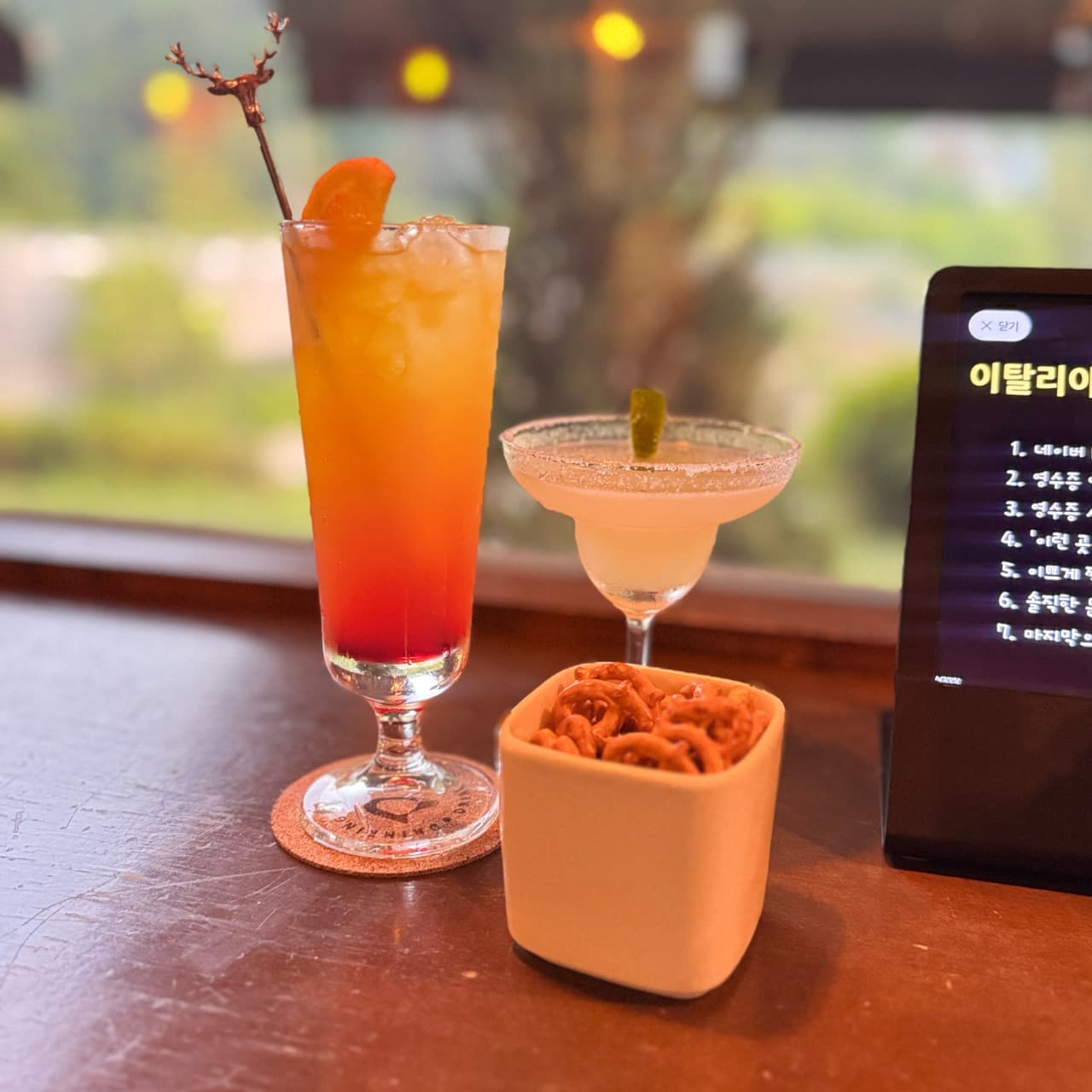 경기도 화성시 드링킹랩 경기 바 1인 다찌석에 놓인 주황빛 칵테일과 소금 묻은 마르가리타. 프레첼 안주와 노란 조명이 비치는 창가 목재 카운터 앞의 혼술 상황.