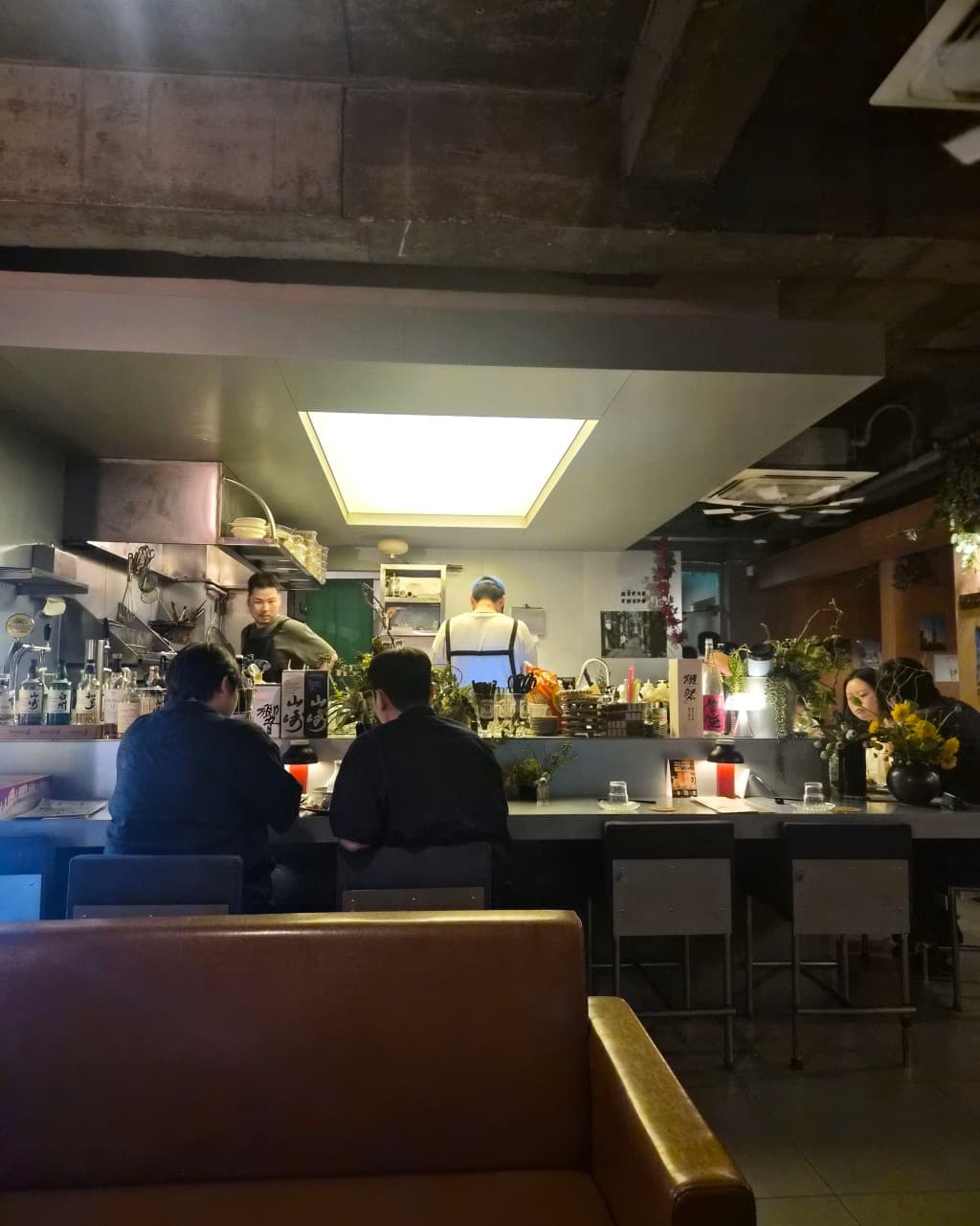 대구광역시 중구 삼덕동 무한궤도 대구의 환한 조명 아래 오픈 주방과 연결된 1인 다찌석에서 혼술을 즐기는 상황. 노출 콘크리트 천장 아래 진열된 주류병과 좌석 전경.