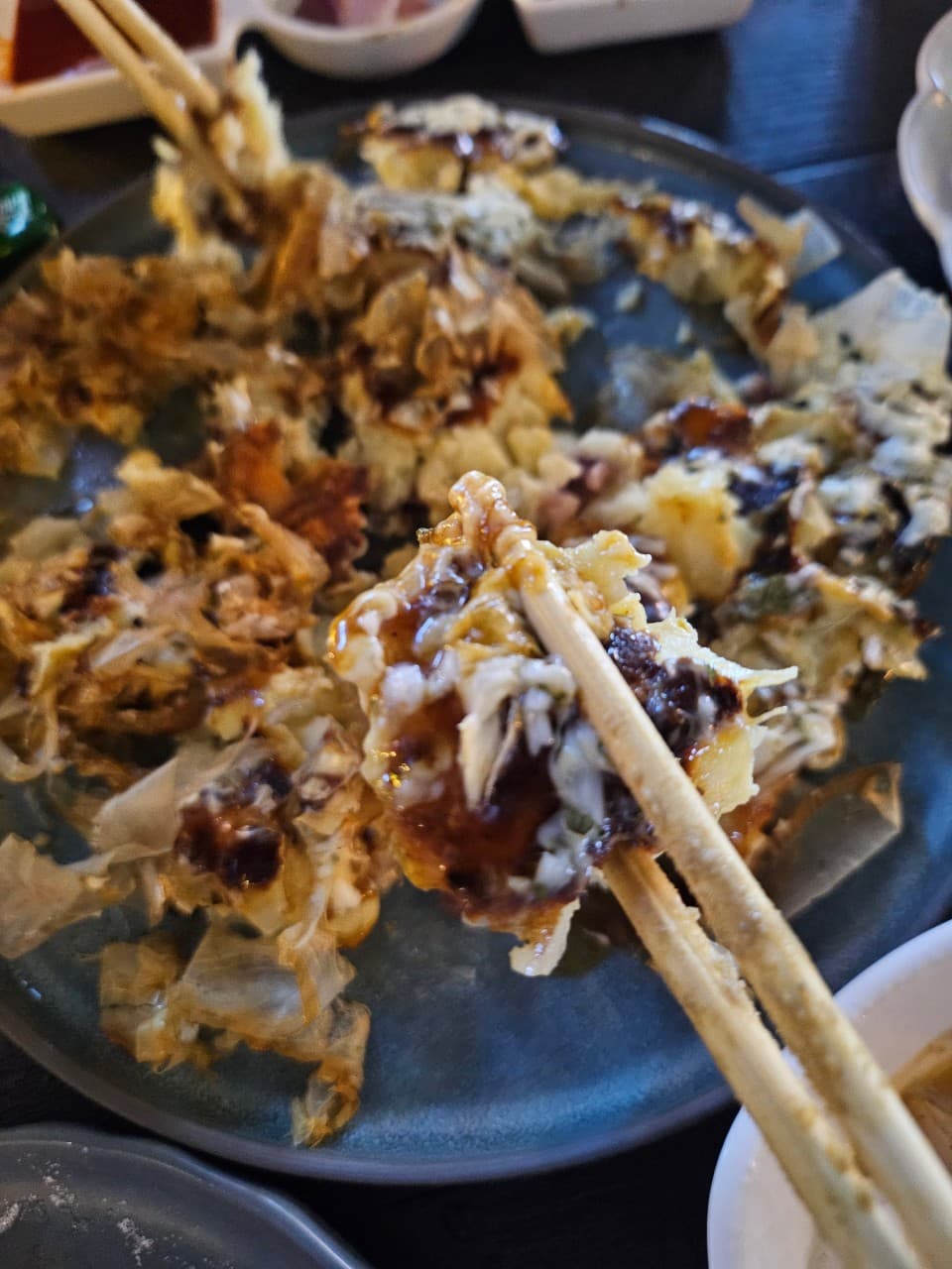 대구광역시 달서구 몽수산 본점 대구 이자카야 혼술 다찌석. 소스가 뿌려진 안주를 젓가락으로 들어 올린 질감과 낮은 조명 아래 1인 식사 상황.