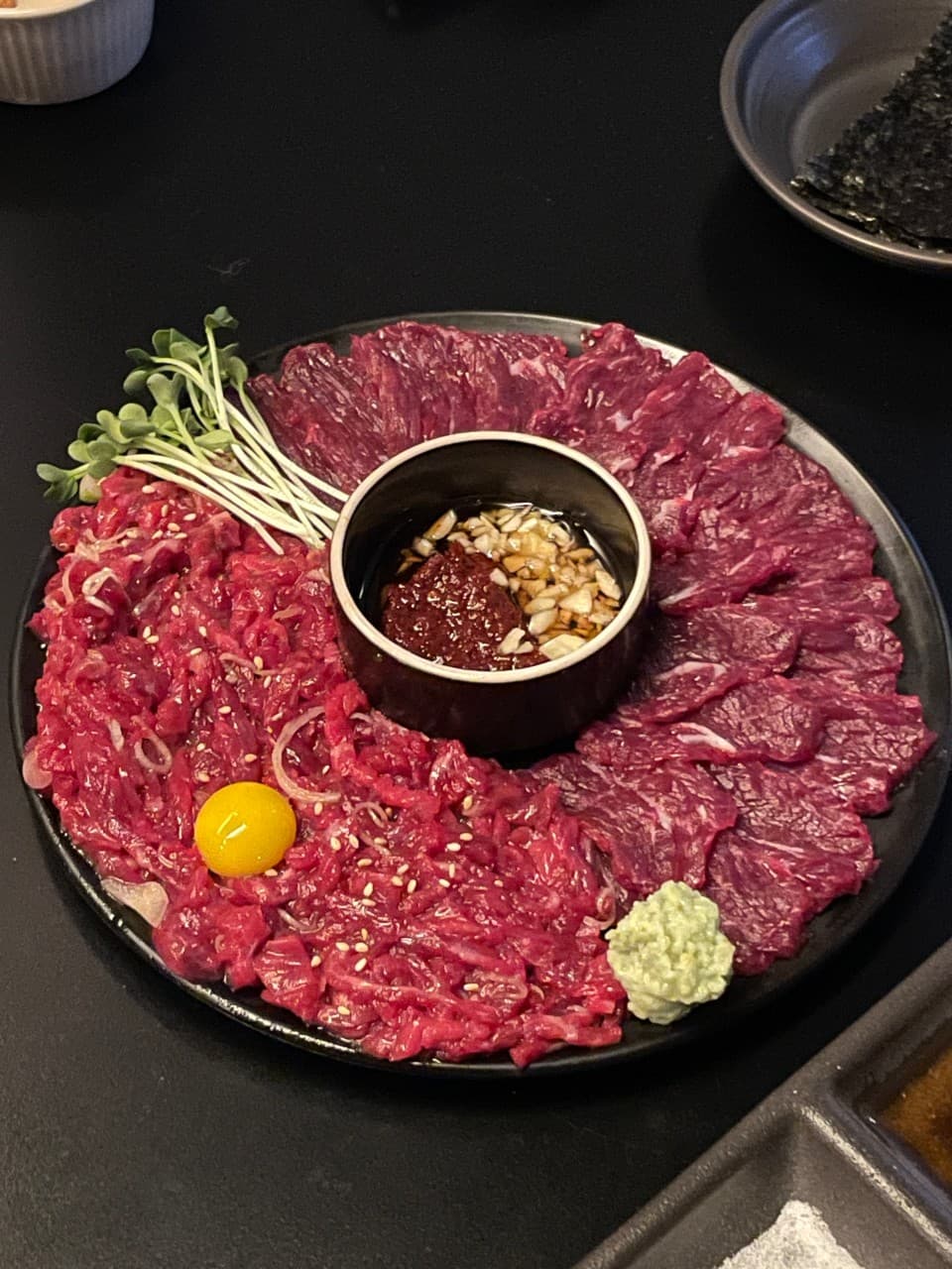 대구광역시 북구 새로나 대구의 기타 요리 혼술 상황. 어두운 조명 아래 다찌석에서 검은 접시에 담긴 붉은 육회와 생고기, 계란 노른자의 질감이 강조됨.