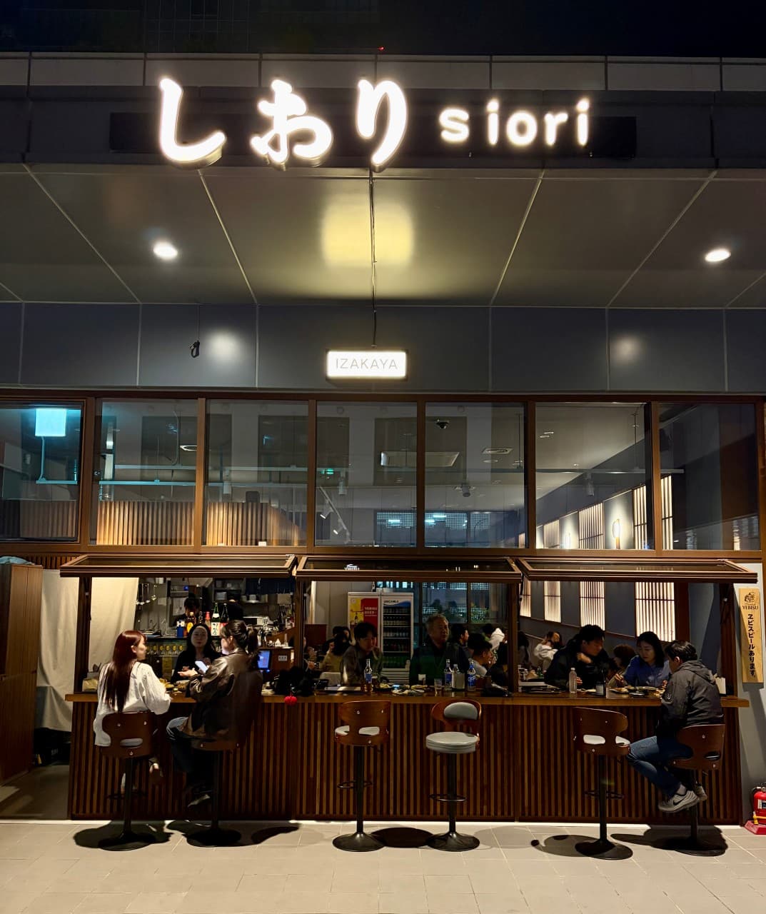 인천광역시 연수구 siori 인천 요리주점 외부. 밝은 조명 아래 개방형 우드 다찌석에 앉아 1인 식사와 혼술을 즐기는 사람들과 테이블 위 술잔이 놓인 상황