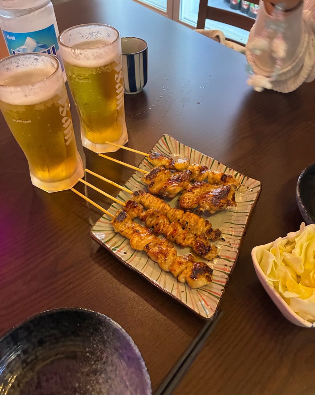 인천광역시 영종구 시마네코 인천 이자카야의 어두운 다찌석 위로 윤기 흐르는 닭꼬치와 생맥주가 놓인 1인 혼술 상황. 조명 아래 정갈하게 차려진 음식의 질감이 돋보임.