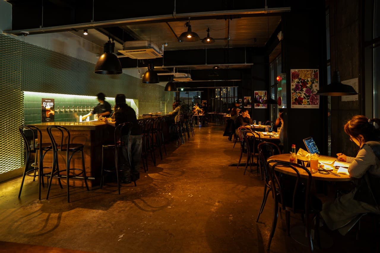 제주특별자치도 제주시 맥파이 탑동점 제주 - 주황색 펜던트 조명 아래 긴 다찌석과 1인 테이블에서 즐기는 혼술. 거품 가득한 수제 맥주를 마시며 개인 작업을 하는 기타 주점