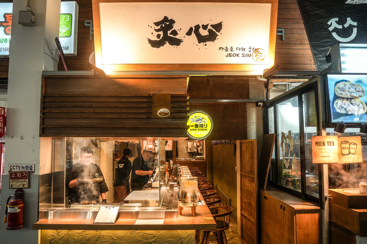 제주특별자치도 서귀포시 적심 제주 이자카야의 노란 조명 아래 일렬로 배치된 다찌석. 연기가 피어오르는 그릴에서 음식을 조리하는 셰프와 혼술을 위한 1인 카운터석이 위치함.