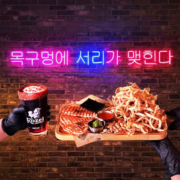 강원특별자치도 춘천시 리얼펍 춘천온의점 맥주 호프. 네온사인 아래 시나몬 가루를 얹은 코젤 흑맥주와 바삭한 먹태 안주. 벽돌 배경의 다찌석에서 즐기는 1인 혼술 상황.