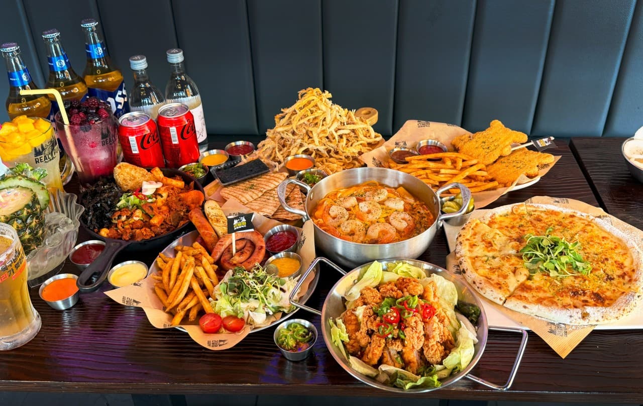 강원특별자치도 춘천시 리얼펍 춘천온의점 맥주 호프 매장. 테이블 위 바삭한 치킨과 피자, 새우 요리가 차려진 모습. 1인 안주와 맥주 병이 놓인 좌석에서 즐기는 혼술 상황을 묘사.