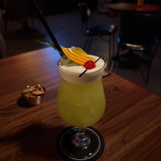 강원특별자치도 속초시 주경야독 바(Bar)의 어두운 조명 아래 나무 테이블 위 거품 층과 오렌지가 올라간 연두색 칵테일과 프레첼. 1인석에서 혼술을 즐기는 상황.