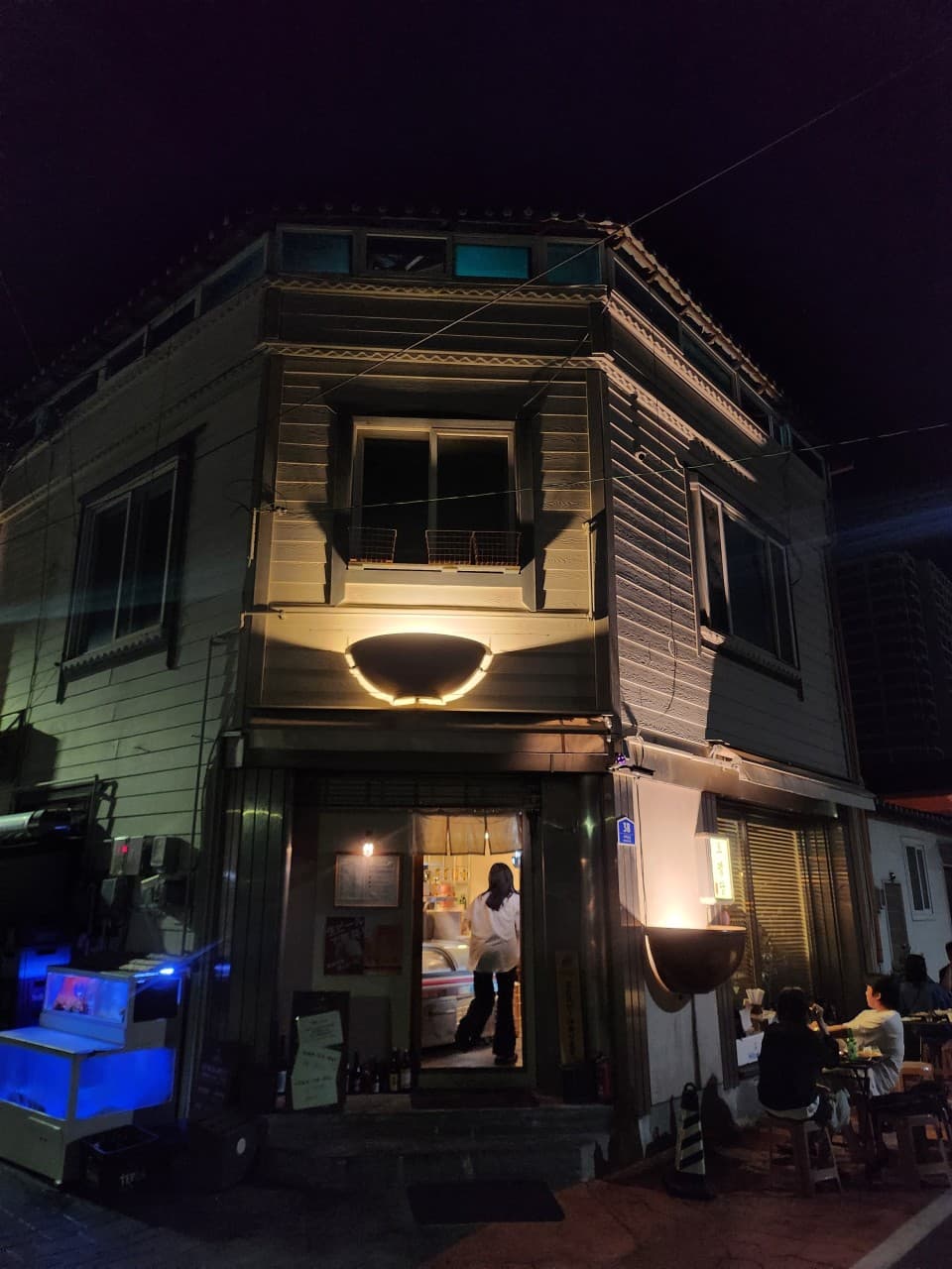 강원특별자치도 속초시 오뎅바 오뎅당의 밤 풍경. 그릇 모양 조명이 비추는 건물 외관과 야외 테이블, 혼술객을 위한 실내 다찌석이 보이는 입구의 모습.