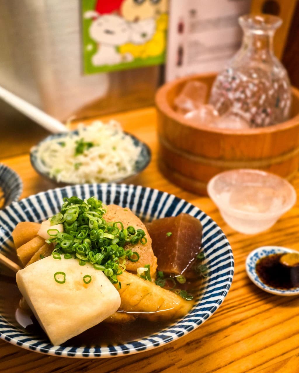 강원특별자치도 속초시 오뎅바 오뎅당. 주황빛 조명 아래 1인 다찌석에 놓인 대파 고명의 오뎅 그릇과 얼음물에 담긴 술병. 혼술 상황의 목재 카운터 테이블 구성.