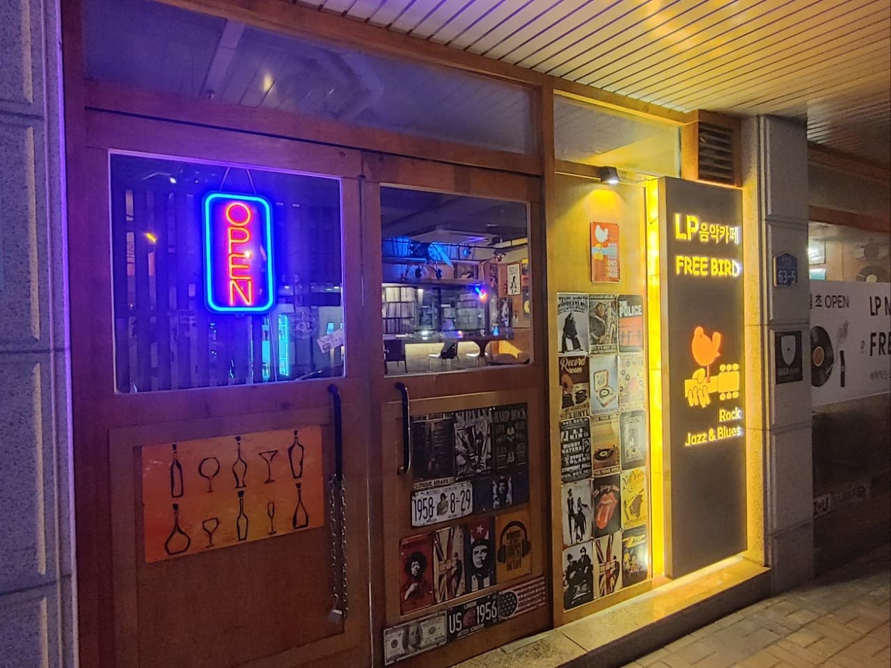 강원특별자치도 원주시 Lp 프리버드 바(Bar) 입구. 네온사인과 포스터가 붙은 나무 문 너머 1인 다찌석과 LP 선반이 보임. 노란 조명 아래 혼술 좌석이 마련된 내부 공간.