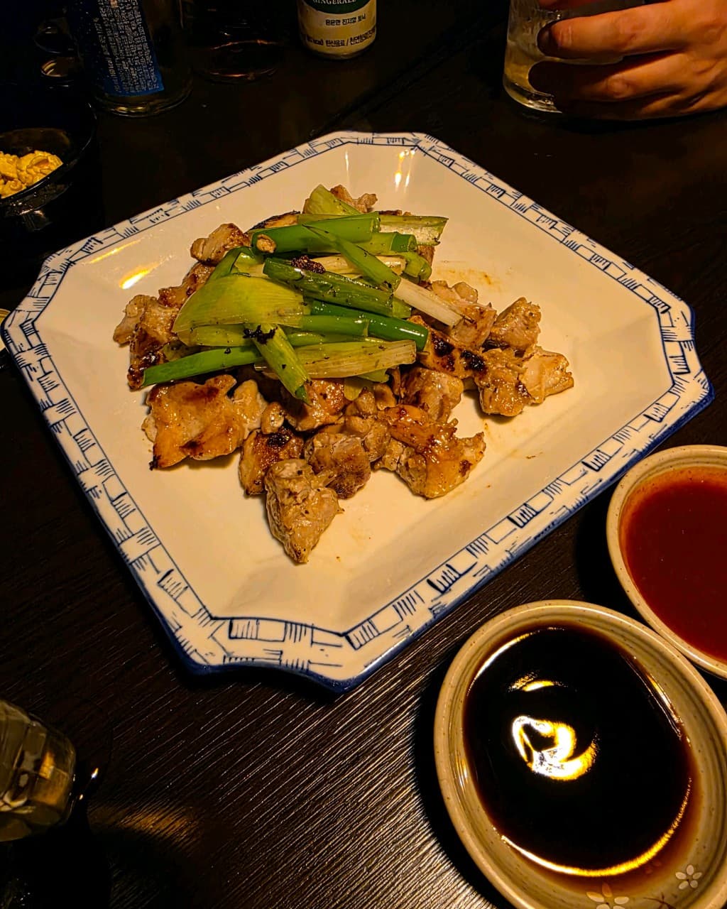 강원특별자치도 강릉시 잉걸불 다찌석 조명 아래 노릇하게 구워진 고기와 대파. 소스 두 종류가 곁들여진 1인 메뉴와 기타 안주를 즐기는 혼술 상황.