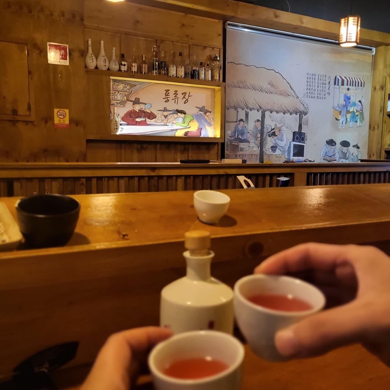 강원특별자치도 춘천시 전통주점 풍류장 - 나무 다찌석에서 붉은색 전통주 잔을 든 모습. 민속화 벽면과 조명 아래 혼술 가능한 카운터 테이블이 배치된 목재 인테리어 공간.