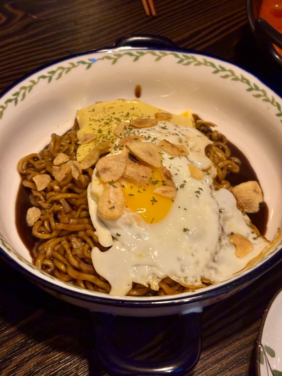 강원특별자치도 춘천시 전통주점 풍류장 다찌석 혼술 - 조명 아래 반숙 계란과 치즈, 마늘 칩이 올라간 짜장라면의 꾸덕한 질감과 나무 테이블