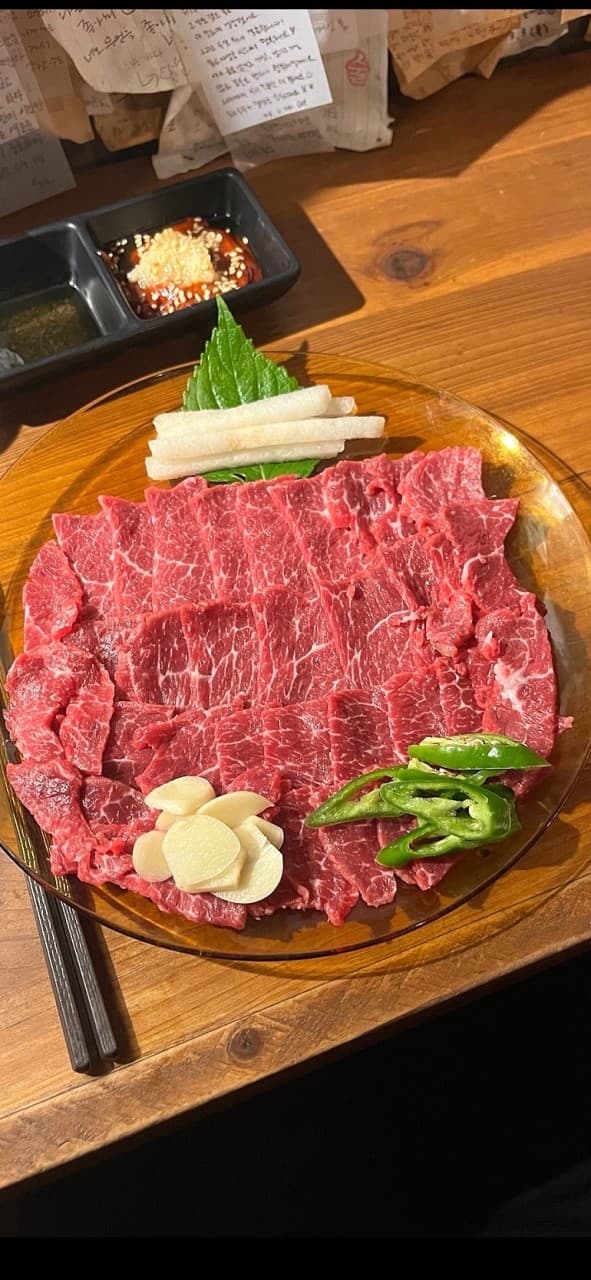 강원특별자치도 속초시 요리주점 아프리카의 다찌석 조명 아래 차려진 1인 생고기 육회와 마늘. 선홍빛 고기 질감이 돋보이는 목재 테이블 위 속초 혼술 상황